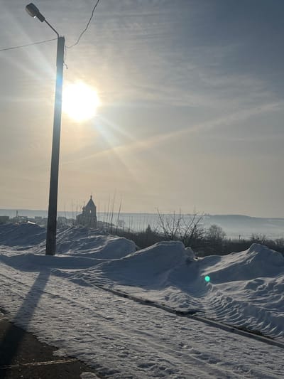 The image displays a bright winter landscape in Kozlovka, Russia, under direct sunlight. A tall, dark utility pole with a streetlamp fixture on top stands on the far left, casting a long shadow diagonally across the snow-covered ground. The lower left foreground features a dark, partially snow-covered paved surface. The middle ground is dominated by extensive, uneven snowdrifts. In the center-left, behind the utility pole and sparse, leafless trees, a light-colored church building with a multi-tiered bell tower and a dome is visible. The background consists of a vast expanse of snow-covered rolling hills under a hazy, bright sky with thin clouds. The sun, a strong light source, is positioned in the upper left quadrant, generating significant lens flare and horizontal light rays across the scene. A distinct bright green lens flare artifact is present within a snowdrift in the lower right area. No individuals, animals, or explicit actions are depicted.
