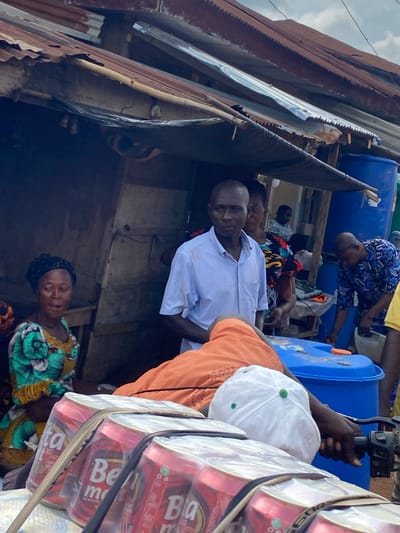 In Amaseri, Nigeria, the image captures a scene outside a rustic, makeshift shelter with a corrugated metal roof. A man wearing a light blue striped shirt stands in the foreground, looking towards the camera. To his left, a woman in a colorful green and yellow patterned dress with a black headscarf is also looking towards the camera, her mouth slightly open. Behind them, other individuals are visible within the shelter. In front of the shelter, a motorcycle is parked, loaded with several packs of red cans, likely beverages, wrapped in plastic. A white baseball cap with green accents sits on the motorcycle seat, next to a person's arm. Large blue plastic barrels are also present in the background. The overall environment suggests a street-side vendor or informal marketplace setting.