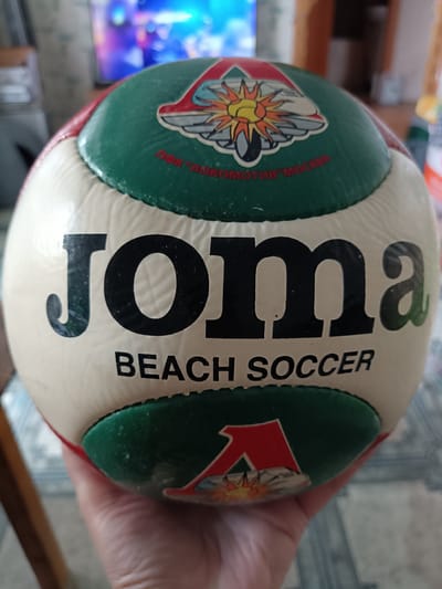 The image shows a close-up of a white and green Joma brand beach soccer ball held in a person's hand. The ball has the Joma logo in large black lettering across its surface. Above the logo, on a green panel, is a circular emblem featuring a stylized sun with rays and what appears to be a train or locomotive, along with the text "ФК "ЛОКОМОТИВ" МОСКВА" (FC "LOCOMOTIVE" MOSCOW). Below the Joma logo, also on a white panel, is the text "BEACH SOCCER". A red and green "A" emblem is visible on the lower green panel of the ball. The background is blurred and shows a television screen displaying colorful imagery and part of a room interior, suggesting the photograph was taken indoors. There is no visible indication of the city of Miasskoye, Russia, within the image.