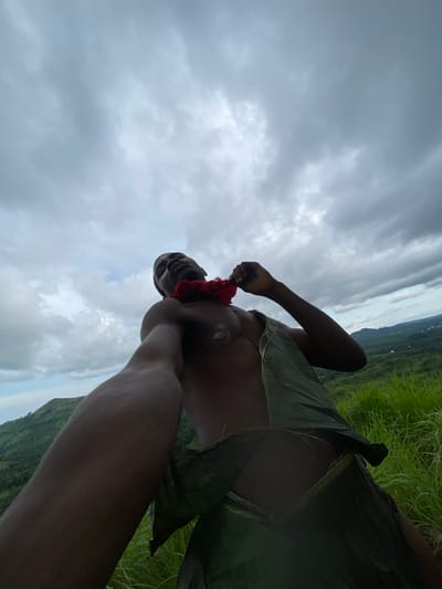 The media file features a low-angle shot of a person outdoors. The subject, who appears to be an adult, is looking upwards and to the left, with their body angled in a way that suggests they are lying down or leaning back. They are wearing a garment made of large, dark green leaves, and a red lei or garland around their neck. Their arms are visible, with one hand near their face. The background is dominated by a cloudy sky, with hints of green hills or mountains in the distance, suggesting a natural, possibly tropical or hilly environment. The overall mood is somewhat dramatic due to the cloudy sky and the upward-looking gaze of the subject. The scene appears to be set in Amaseri, Nigeria. There is no visible text in the image.