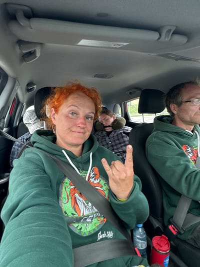 The image shows the interior of a car with four occupants. In the foreground, a woman with bright red hair is in the driver's seat, facing the camera and making a hand gesture. She is wearing a green hoodie with a colorful graphic. To her right, a man with glasses is driving the car. In the back seat, a person is asleep, leaning on a travel pillow, and another person is partially visible behind the sleeping individual, wearing a grey cap. The environment appears to be a car interior on a cloudy day. There is no information in the image that can definitively place the location in Tatarstan, Russia.