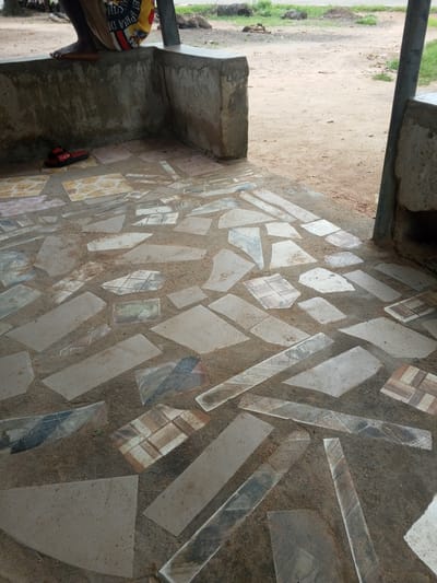 The image shows a close-up view of a mosaic tiled floor, likely a patio or walkway. The tiles are irregular in shape and size, with a variety of colors and patterns, including plain beige, grey, and some with intricate designs resembling wood or marble. They are embedded in a rough, sandy grout. To the left of the tiled area, a person's lower legs and feet are visible, with one foot resting on a low concrete wall. The person is wearing a colorful patterned cloth around their waist. On the ground near the wall are a pair of red sandals. The setting appears to be outdoors, with a sandy, dirt ground visible in the background. The lighting suggests it is daytime, possibly morning or afternoon, with shadows indicating sunlight. The overall impression is of a rustic, possibly unfinished outdoor space in a rural or semi-rural location in Gindin Dutse, Nigeria. There is no visible text in the image.