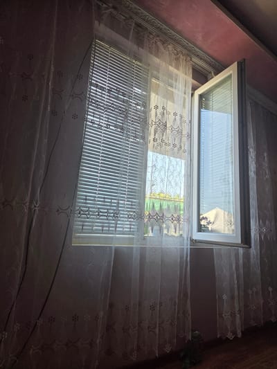 A partially open window with white blinds and white lace curtains is depicted in a room. The window is framed by a white structure, and outside, greenery and a light-colored structure are visible. The room has a pink ceiling with decorative molding and appears to be lit by natural light coming through the window. The floor is made of wood. The scene is set in a domestic interior in Todor Ikonomovo, Bulgaria.