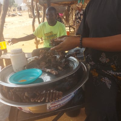 A man in a green t-shirt stands near a circular tray of cooked meat and ribs. A woman with a beaded bracelet reaches into the tray with her hand, likely serving the meat. A small blue plate rests on top of a white container within the tray. The setting appears to be outdoors, possibly a food stall or market. The image is somewhat blurry and taken from a slightly high angle, with a shallow depth of field.
