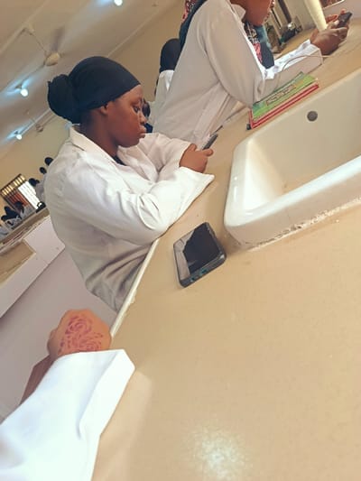 This image captures an indoor scene within a classroom or laboratory environment in Rafin Pa, Nigeria. The perspective is a first-person view, with a human hand in the lower-left foreground displaying a red/brown floral (rose-like) henna design on the back, and wearing a white long-sleeved garment.
To the right of the foreground hand, a dark-colored smartphone with a visible screen reflection and charging port is resting face-up on a light-colored, possibly beige or off-white, laboratory table. Further along the table, a young woman, likely a student, is seated. She wears a white lab coat over a lighter garment and a black head covering, with her hair gathered underneath. Her gaze is directed downwards as she holds and interacts with a dark-colored smartphone with both hands.
Adjacent to this woman, another individual is seated, also wearing a white lab coat and a patterned head covering. Their face is partially obscured. This person is holding another dark-colored smartphone. Next to them on the table is a stack of several books, featuring colorful covers primarily in red and green hues, positioned near a white porcelain laboratory sink with a visible drain.
In the background, multiple other individuals wearing white lab coats are visible seated at similar tables, suggesting a group setting. The room features multiple circular ceiling light fixtures and at least one ceiling fan. Light-colored cabinets or shelving units are visible along the far wall on the left.