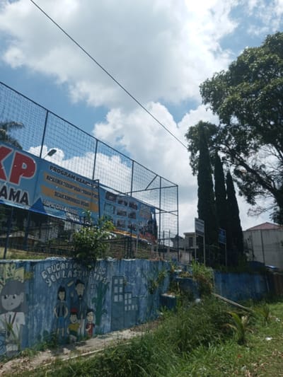 A slightly low-angle shot depicts a public space in Kioumaknao 2, Laos. A blue fence topped with netting separates the viewer from an elevated area containing a large banner. The banner has text in an unidentified language (possibly Indonesian or a similar regional language), advertising programs such as "MANAJEMEN SERKANTORAN LAYAKAN PISNIS" and "DESAIK KOMUNIKASI VISUAL". Below the fence sits a blue wall, adorned with a mural depicting stylized figures (including children and an adult) and the English text "EDUCATION GIVES YOU LASTING POWER". Foreground is dominated by overgrown vegetation. Beyond the fence, other buildings and trees are visible. The sky is partly cloudy.