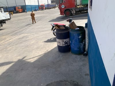The image displays an outdoor industrial yard in Babawa, Nigeria, under bright daylight conditions, characterized by long shadows cast on the concrete or asphalt ground. In the mid-ground, a lone male figure, dressed in light brown attire, walks away from the camera, facing the distant background. To the right, two blue barrels, appearing repurposed as refuse containers, stand against a building wall painted white above and dark blue below; both barrels contain cardboard and other visible waste. A red motorcycle is partially visible behind the barrels. The background features a large, light-colored building with horizontal blue banding, likely a warehouse or industrial structure. Multiple large trucks, including blue cargo trucks and red semi-trailer cabs, are distributed across the expansive yard. Several smaller figures are visible near the trucks in the far distance, alongside a small blue car.