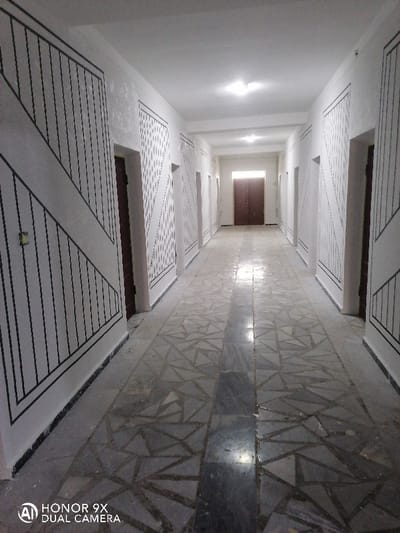 The image displays an empty, interior hallway located in Samarkand, Uzbekistan. The perspective is from the nearer end, extending down the corridor towards a pair of closed, dark brown double doors positioned centrally at the far end. White walls line both sides of the hallway, adorned with a repetitive decorative pattern of dark, thin geometric lines creating vertical and diagonal segments within rectangular frames. Multiple dark brown doors are recessed into these patterned walls along the length of the corridor. The floor is tiled with a mosaic-like pattern of light and dark grey irregular geometric shapes, featuring a prominent darker grey or black solid strip running down the center longitudinally. The white ceiling contains at least two square, flush-mounted overhead light fixtures providing illumination, with their reflections visible on the polished floor tiles. No individuals, actions, or movable objects are present in the scene.
