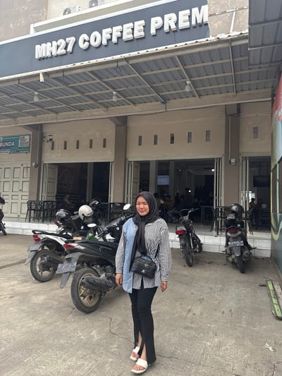 A woman wearing a black hijab and a striped shirt is standing on a concrete sidewalk in front of a building. The building has a sign above the entrance that reads "MH27 COFFEE PREM". There are several motorcycles parked on the sidewalk. The woman is looking to her left and smiling. The environment appears to be a commercial area in Meunasahmanyet, Indonesia. The image is taken from a medium distance.