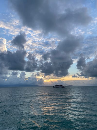 The image shows a body of water, likely the sea, with a passenger ferry on the right side. On the horizon, there is a landmass. The sky is filled with clouds, with some areas of clear blue sky visible. The setting sun creates a golden glow on the water and through gaps in the clouds. The location is possibly near Thong Nian, Thailand, given the likely geographical features. There is no evidence of people, actions or interactions.