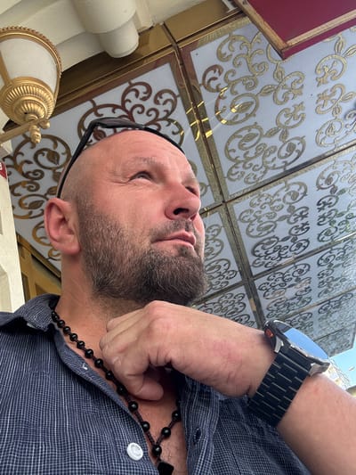 The image is a close-up, low-angle shot of a bald, middle-aged man with a beard, looking upwards and to the right. He is wearing a dark blue checkered shirt and a black beaded necklace. A hand is resting on his chin. On his left wrist, he wears a black metal watch with a blue-toned digital display. In the background, there is a decorative ceiling with gold patterns and a golden light fixture on the left. The man's eyeglasses are perched on top of his head. The scene appears to be outdoors, possibly in a cafe or sheltered seating area, with the ornate ceiling suggesting an establishment with a distinct architectural style. There are no explicit indicators to place this scene specifically in Avcılar, Turkey, or any other city.