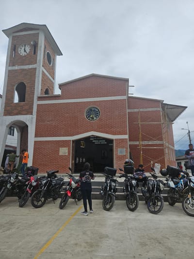 This image captures a wide shot of a brick building with a prominent clock tower in Bucarasica, Colombia, under an overcast sky. The building, likely a church or public structure, features red brick construction with light grey concrete accents.

On the left, a multi-story clock tower stands, exhibiting arched openings on its lower level and a large circular clock face on its upper section, indicating approximately 11:30. A white public address speaker is mounted to the right of the clock face. An oval-shaped window is visible on the right facade of the tower's upper section.

The main section of the building, immediately to the right of the tower, presents a large brick facade with a central arched entrance that leads into a dimly lit interior. Above the entrance, a circular stained-glass window depicts a figure, possibly religious. Yellow scaffolding is erected against the right side of the main building, extending upward, indicating ongoing construction or renovation.

In the foreground, a concrete plaza serves as a parking area for approximately eight to ten motorcycles, predominantly dark-colored, with some featuring red or blue accents. Most motorcycles are equipped with black rear storage boxes. A yellow line is painted on the concrete surface.

Multiple individuals are present. One woman stands with her back to the camera, positioned on the right side of the motorcycles, wearing a black shirt with "YUS" printed on the back and dark jeans. Several men are scattered around the scene; one is seated on the steps leading to the building entrance, while others stand near the motorcycles or by the left archway of the clock tower. One man on the far right wears a black and red patterned shirt. Two men are visible near the base of the clock tower, one in an orange shirt appears to be looking at a phone.

In the background, behind the building to the right, utility poles with wires are visible against a backdrop of distant hills or mountains. The sky is uniformly grey and cloudy.