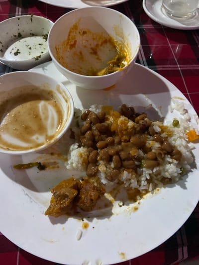 The image displays a close-up, top-down view of a partially consumed meal served on a checkered tablecloth, likely indoors under artificial lighting. The central subject is a large white plate holding a substantial portion of white rice. The rice is heavily topped with brown beans and a light brown, thick sauce, with scattered small pieces of orange vegetables (possibly carrots) and green peas visible throughout. To the right of the rice and beans, there are several chunks of what appears to be stewed meat, dark brown and tender, resting in a rich, savory sauce.
Surrounding the main plate are several small, white ceramic bowls containing remnants of various dishes. One bowl to the upper left holds a creamy, yellowish-green sauce or soup, possibly a curry, with some solid components still visible. Below it, another small bowl is largely empty, with sparse traces of green herbs or spices clinging to its interior. To the lower right, a third small bowl contains a thicker, light brown, frothy liquid, resembling a lentil soup or another type of sauce.
The dishes are arranged on a red and black checkered tablecloth. The lighting suggests an overhead source, casting distinct shadows and creating a somewhat dark, ambient atmosphere, implying the meal is taking place in the evening or at night. The informal presentation of the food and table setting points to a casual dining experience, possibly in a local eatery or home in Bagamoyo, Tanzania, given the typical East African cuisine featuring rice, beans, and stew. No people are visible, but the state of the food indicates active consumption or a recently finished meal. No text is visible in the image.
