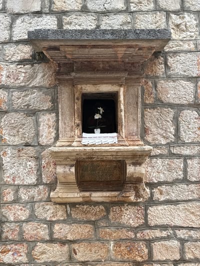 A small, ornate stone niche is set into a rough-hewn stone wall, characteristic of the architecture in the old town of Grad Stari Grad, Croatia. The niche features a small white orchid in a white pot placed on a white crocheted cloth. Above the niche, a dark, textured stone slab juts out, supported by carved stone brackets. Below the niche is a weathered bronze plaque with engraved text. The overall scene suggests a devotional space integrated into the historic urban fabric.