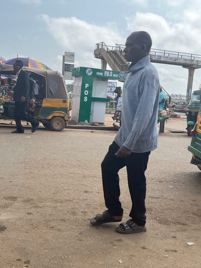 A man in a light blue sweater and black pants walks on a dusty road in Gura Top, Nigeria. He is wearing black sandals. To his left, a yellow and brown auto-rickshaw is parked. Behind the auto-rickshaw, a sign for "TotalEnergies" is visible, along with fuel prices. To the right of the auto-rickshaw, a green kiosk with "P.O.S OUTLET" signage is present. In the background, a pedestrian overpass spans the road. The sky is overcast.