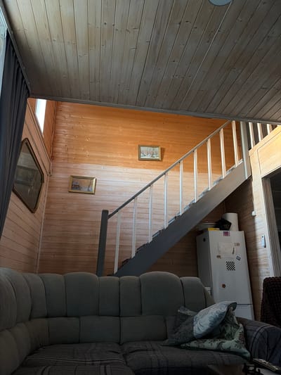 This is an indoor, low-angle shot of a room with wooden walls and ceiling. In the foreground, a large, sectional sofa is visible, covered with throws and pillows. Behind the sofa, a staircase with white spindles and a grey railing leads upwards. To the right of the staircase, a white refrigerator is situated against the wooden wall. A roll of paper towels and a black trash bag are on top of the refrigerator. On the wooden wall to the left of the staircase, two framed landscape paintings are hung. Further to the left, a large, ornate mirror is mounted on the wall. The room is lit by natural light coming from an unseen window. This is a domestic scene located in Проводник, Russia.