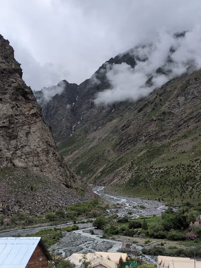 The image depicts a panoramic view of a valley in Gemur, India, under a cloudy sky. Towering, rugged mountains flank the valley, with the left side dominated by a steep, rocky cliff face and the right side showcasing gentler, green slopes dotted with vegetation. A winding river, characterized by its rocky bed and clear, flowing water, meanders through the valley floor. Patches of fog or clouds are visible clinging to the mountain peaks and slopes, adding a dramatic effect to the landscape. In the foreground, a portion of a building with a corrugated metal roof is visible on the left, and several tents, likely part of a campsite, are spread out near the riverbank on the right. The scene conveys a sense of natural grandeur and a remote, serene environment.
