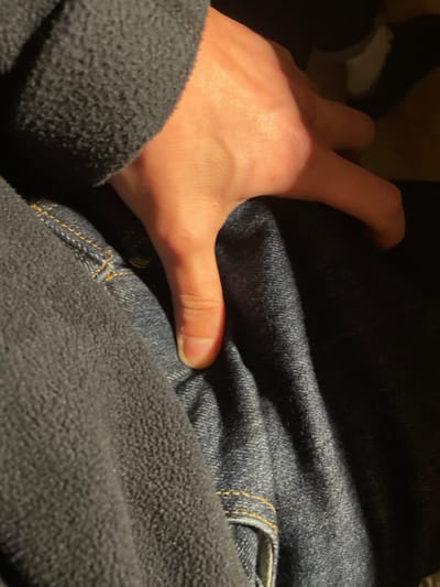 A dimly lit, close-up image features a fair-skinned human hand grasping a section of dark blue denim fabric, likely from a pair of jeans. The hand emerges from behind a dark grey or black fleece material, possibly a sleeve or another garment, which covers the lower portion of the arm and part of the denim.
The hand, illuminated by a direct light source, presses its thumb into the denim, causing a visible indentation. Visible details of the hand include fine lines, subtle veins, and a short, clean fingernail on the thumb. The dark blue denim displays its characteristic woven texture and features yellow-orange stitching, typical of jeans, near what appears to be a pocket seam on the right side of the frame. The fleece fabric has a soft, textured surface.
The scene suggests an indoor setting or nighttime due to the focused lighting and the surrounding shadows. The image primarily highlights the textures of the skin, denim, and fleece, and the tactile interaction between the hand and the fabrics. No specific activity beyond the act of gripping is discernible, and no visible text is present. The provided context for the location is Ganei Tikva, Israel.