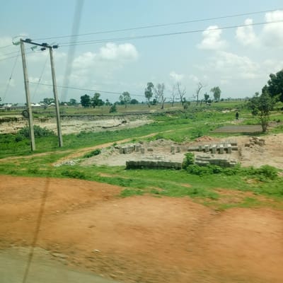 The image captures a rural landscape in Mutum Biyu, Nigeria, characterized by a light blue sky with scattered white clouds. The foreground is dominated by an orange-brown dirt road, blurred by motion, which transitions into a grassy area. Beyond the grass, a shallow, murky watercourse is visible, bordered by eroded, sandy banks. Construction materials, specifically stacks of concrete blocks, are present in a cleared area on the right side of the watercourse. Further in the background, several residential buildings with light-colored roofs are situated amidst a scattering of green trees and vegetation. A power line supported by two wooden poles runs horizontally across the mid-ground, with thick black wires extending from them. A single figure, dressed in dark clothing, is walking in the distance on the right side of the frame, near a flat, patterned surface that appears to be drying bricks or tiles. The overall scene depicts a developing rural area with ongoing construction and basic infrastructure.