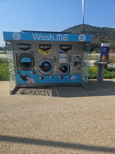The image displays an outdoor, self-service laundry facility, branded "Wash.ME," located under a clear blue sky in Fréjus, France. The facility consists of a gray metal structure with a prominent blue header featuring the "Wash.ME" logo, a steaming shirt icon, and a "20kg" basket icon. It contains three front-loading machines: one "SÈCHE 20 KG" (dryer) priced at 3€ on the left, and two "LAVE" (washing machines)—a "20 KG" model for 12€ in the center, and a "9 KG" model for 5€ on the right. The central washing machine features a yellow "Nouveau! LAVE ET SÈCHE TOUT EN UN" sticker. Informational graphics are present below the machines; specifically, a smiling woman with hair flowing upwards is depicted on the dryer's lower panel, and a pile of white laundry is shown below the 9kg washer. A central payment and control interface is situated between the two washing machines. The structure stands on a light tan gravel surface, with distinct shadows indicating direct sunlight. Green vegetation, interspersed with small yellow flowers, flanks the gravel area. To the immediate right, a blue "VINCI Autoroutes Collecte sélective" recycling sign and a light gray concrete stand are visible. In the background, a road is present, beyond which a large, densely tree-covered hill or mountain rises under the clear sky. No individuals are present, and no actions or interactions are occurring within the scene.