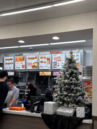 A fast-food restaurant's menu board is displayed in vertical format. It features pictures of various burgers, chicken sandwiches, shakes, and sides. A small, decorated Christmas tree sits on the counter in front of the menu, along with gift-wrapped boxes. Two people are standing at the counter.