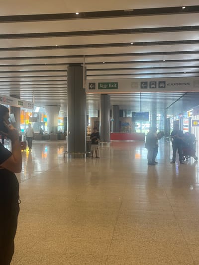 This is a vertical, wide shot of a brightly lit interior, likely an airport or train station. The setting is modern with a polished, speckled floor that reflects the overhead lighting and the architecture. The ceiling features a series of parallel, illuminated white panels running the length of the space.
In the foreground, a person's arm and shoulder are visible, as they appear to be reclining or sitting on something out of frame. Their clothing is dark.
Further into the scene, several people are visible, some walking with luggage, others standing or sitting. One group in the background appears to be waiting. The people are diverse in their attire and activities.
There are several signs with text and icons visible. One prominent sign, in the left mid-ground, has the text "EXPRESS" vertically oriented, along with icons for a bus and a car, and arrows. Below this, another sign reads "tlp Exit" in green. Another sign further back indicates "Parking".
The lighting suggests it is daytime, with bright interior lights and natural light filtering in from large windows or openings in the distance, creating reflections on the floor. The overall atmosphere is one of transit and movement typical of a transportation hub. The scene appears to be set in Parakar, Armenia.