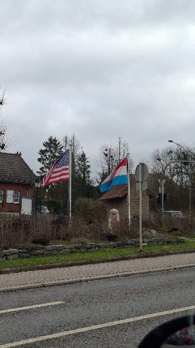 The scene depicts an outdoor roadside view in Frisange, Luxembourg, under an overcast sky. In the midground, two national flags are prominently displayed on separate silver poles: an American flag (Stars and Stripes) on the left and a Luxembourgish flag (red, white, and light blue horizontal stripes) on the right.
To the left of the American flag, a section of a two-story building with a reddish-brown tiled roof and light brown stucco walls is visible, featuring white-framed windows with partially open white shutters. A black wall-mounted lantern is affixed to this building. Further back, dark green coniferous trees are present. Behind the Luxembourgish flag, a smaller stone structure with a similar tiled roof is situated. To the right of this structure, a stand of deciduous trees with bare branches and several visible nests is present.
Between the Luxembourgish flag pole and the small stone building, a decorative cylindrical marker, painted white with red and black accents, stands on a small mound. A round, grey road sign on a pole is positioned to the right of the Luxembourgish flag. A black street light with a curved arm is visible in the upper right.
The foreground consists of a grey asphalt road with parallel solid and broken white lane markings. A concrete curb separates the road from a grey paved sidewalk. Beyond the sidewalk, a strip of green grass borders a low dry-stone wall. Behind this wall, an area of dry, brown vegetation extends across the midground. The lower right corner of the image shows a partial reflection of a vehicle's dashboard, indicating the photo was taken from inside a car. No people or active interactions are visible.