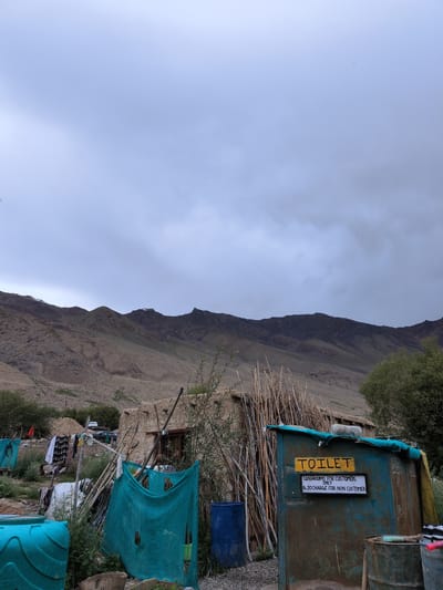 A rustic, weather-beaten toilet structure stands in a remote, mountainous area, possibly near Khardung, India. The structure is painted a faded teal and features a yellow sign that reads "TOILET". Below this, a smaller white sign states "WASHROOMS FOR CUSTOMERS ONLY Rs. 20 CHARGE FOR NON CUSTOMERS". The toilet is surrounded by a natural, arid landscape with rocky, barren hills in the background. Debris and makeshift structures, including poles and tarps, are scattered around the scene, suggesting a basic, temporary settlement or service area. A large blue plastic tank is visible to the left of the toilet, and a dark blue barrel is to its left. The sky is overcast with heavy clouds.