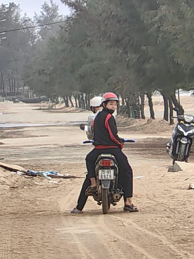 The image shows two individuals on a stationary silver motorbike on an unpaved sandy road in Thuận An Ward, Vietnam. The person in the foreground, identified as the driver, wears a red helmet, a black long-sleeved top with red accents, and black pants, and is looking directly at the viewer. Their feet, clad in dark sandals, are on the ground, supporting the vehicle. A second individual, wearing a white helmet and a light-colored, patterned top, is visible as a passenger behind the driver. The motorbike bears Vietnamese license plate "75-H1 6929" and features blue handlebar grips. To the right, a black and silver motorbike is partially visible, also stationary on the sandy terrain. The background is dominated by a dense line of evergreen trees. Further in the distance, behind the trees, a dark-colored boat or vessel is visible. The sandy ground shows multiple tire tracks, and a pile of debris, including blue fabric, is present on the left side of the frame. The sky is overcast.