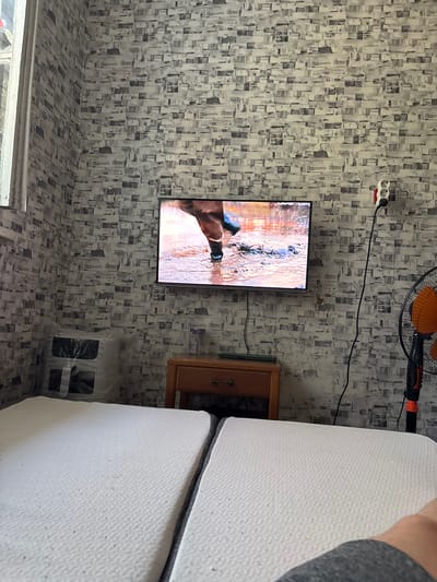 The image shows a room with a television screen displaying a close-up of a horse's leg wading through water. The walls are covered in a grey and white patterned wallpaper. To the left of the television, there is a window with white trim. Below the television, there is a wooden nightstand with a drawer. To the right of the nightstand, a black electrical cord snakes up from the floor to a white electrical outlet with three sockets, and further to the right, a black floor fan with an orange grille is visible. In the foreground, two white mattresses are laid out, suggesting a makeshift sleeping area. A large, clear plastic bag containing dark objects is situated on the left side of the room, near the mattresses. A small purple ribbon is tied around a clear glass object on the nightstand. The scene appears to be in a room in Tarasovsky, Russia.
