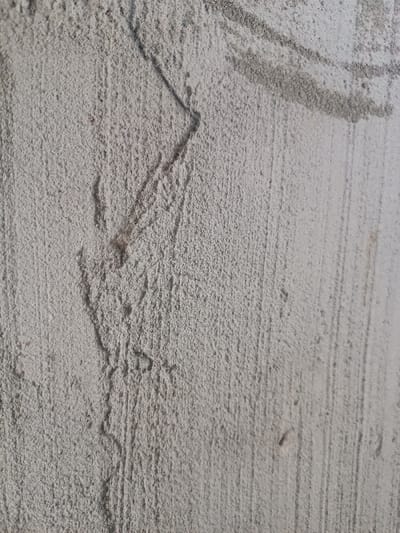 The image displays a close-up view of a light gray, textured wall surface located in Elatan, Uzbekistan. The surface exhibits a rough, granular finish, consistent with a plastered or rendered wall. Prominent features include several dark, thin, linear marks: an angular, V-shaped mark in the upper left quadrant, and a wider, curved mark in the upper right. Vertical striations, possibly from a trowel or finishing tool, are visible across the entire surface, particularly on the right side. No individuals, discernible objects other than the wall itself, or specific actions are depicted within the frame.