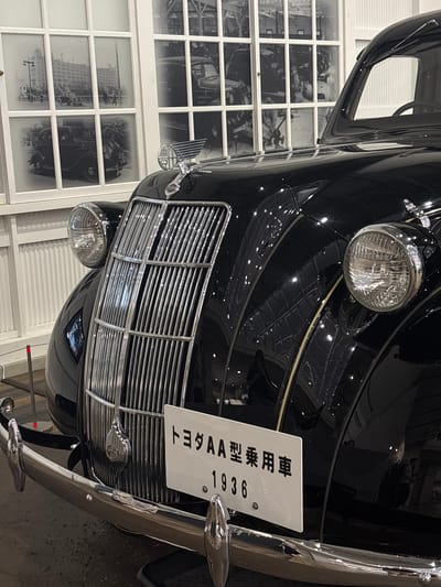 The image shows the front of a black vintage Toyota AA passenger car from 1936, displayed at the Toyota Commemorative Museum of Industry and Technology in Nagoya, Japan. The car features a prominent chrome grille, two round headlights, and a hood ornament. A license plate with Japanese text and "1936" is attached to the front bumper. Behind the car, a wall with large windows displays historical black and white photographs of early automobiles and factory scenes, offering a glimpse into automotive history. The environment appears to be an indoor exhibition space with controlled lighting, highlighting the car's polished surface.
