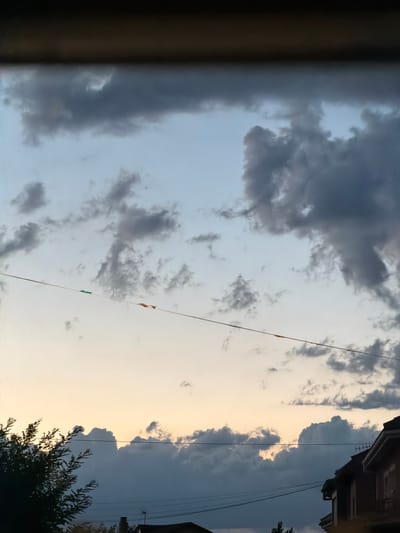 The image depicts a view of the sky from what appears to be a residential area. The sky is filled with a mix of light blue and soft yellow hues, indicative of either early morning or late evening. Dark gray clouds are scattered across the sky, with some appearing more dense and voluminous than others. A thin, taut wire stretches horizontally across the frame, with small, colorful pieces of material attached to it at intervals. In the foreground, the dark silhouette of trees and rooftops of houses is visible against the lighter sky. The architecture of the buildings suggests a European setting, and specifically within the city of Arcahueja, Spain, the presence of residential structures and overhead utility lines is common. There are no people or discernible actions taking place in the image; it is a static scene capturing a moment in time.