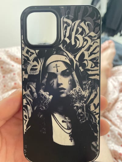 The image shows a close-up of a person's hand holding a black smartphone case. The case features a black and white graphic of a tattooed nun with a cross on her forehead and a rosary around her neck. The background is a blurred pink and white patterned fabric, possibly a bedsheet. In the far background, a person's legs are visible, indicating they are lying down. There is no indication of the city of Paczkowo, Poland, in this image.