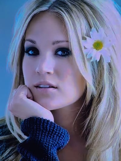 The media file features a close-up, tilted shot of a young woman with blonde hair adorned with a white daisy. She is wearing a dark, textured garment, possibly a sweater or a thick scarf, around her shoulder and neck. Her left hand is resting on her chin, and her eyes are open, looking towards the viewer. The lighting is soft, with a cool tone, suggesting an indoor setting or possibly a subdued outdoor environment. The overall mood is serene and contemplative. The background is out of focus, but the color palette suggests a predominantly blue and light purple hue, creating a dreamy and ethereal atmosphere. There is no visible text in the image.