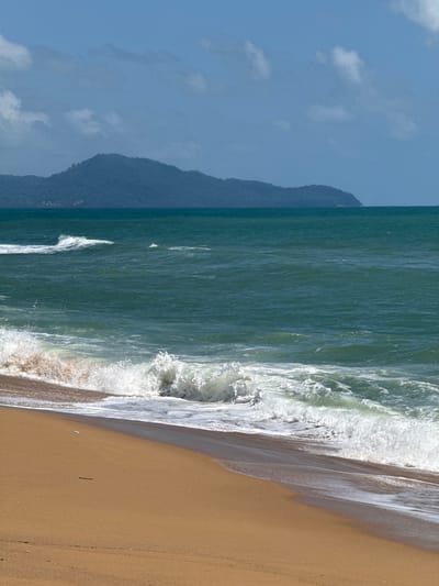 The image shows a tropical beach scene with a wide expanse of sandy shore in the foreground, sloping down to the turquoise ocean. Waves are breaking on the shore, creating white foam and spray. In the distance, a lush green, mountainous island with a hazy outline rises from the sea, under a bright blue sky dotted with wispy clouds. The overall impression is one of natural beauty and tranquility.
While the image depicts a generic tropical beach, the specific location mentioned, Ban Tha Chat Chai, Thailand, is known for its scenic coastline and proximity to islands. The presence of a mountainous island in the distance is consistent with the geography of this region, which is part of the Andaman coast.