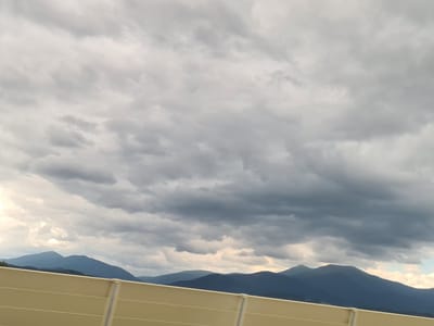 The image captures a panoramic view of a mountain range under a dramatic, cloudy sky. The mountains, rendered in deep blues and purples, are silhouetted against the lighter tones of the clouds, which range from bright white near the horizon to dark, heavy gray overhead. A tan-colored barrier, possibly a sound barrier or guardrail, runs horizontally across the bottom of the frame, partially obscuring the lower slopes of the mountains. The sky is filled with a complex pattern of clouds, suggesting an overcast or potentially stormy day. No people or animals are visible in the image. The scene appears to be a natural landscape with man-made elements in the foreground, likely a roadside view. The location is identified as Pausendorf, Austria.