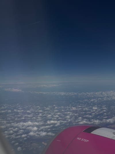 The image is an aerial view from an airplane window, looking out at the sky and clouds. The top portion of the image is a deep blue sky with a faint contrail from another aircraft. Below the deep blue sky, there are layers of lighter blue sky with wispy clouds. The bottom portion of the image shows a dense layer of white, fluffy clouds covering the ground, which is only faintly visible through some gaps in the clouds. A portion of the airplane's wing, painted in a bright magenta color, is visible in the lower right corner of the image. White text that reads "NO STEP" is printed on the wing. There are no people or specific landmarks visible to indicate the city of Chernik, Bulgaria.