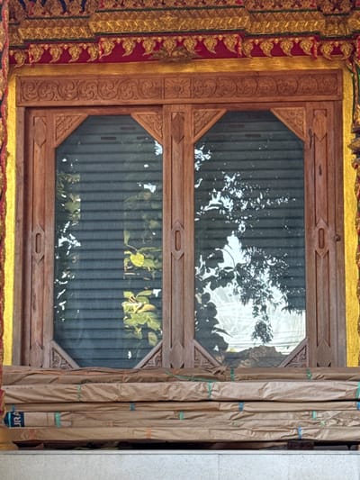 This image shows the exterior of a building in Baan Tha Po, Thailand, featuring a wooden window with intricate carvings. The window is divided into two panels, each with a mirrored surface reflecting trees and foliage. The window is framed by ornate, gold and red decorative elements characteristic of Thai architecture. Below the window, several items are stacked and wrapped in brown paper, secured with colored strings. The overall impression is one of traditional craftsmanship and a serene, possibly temple-like, setting. There are no people visible in the image.