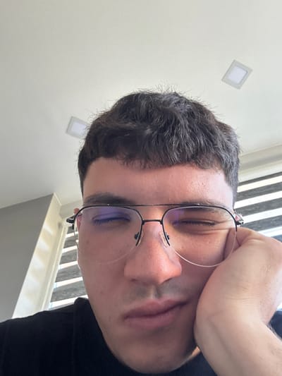The image is a close-up, low-angle selfie of a young man with dark hair, wearing glasses and a black shirt. He is resting his head on his hand, with his eyes closed and mouth slightly open, suggesting he is tired or sleepy. The background shows a white ceiling with two square lights and a window with horizontal blinds. The scene appears to be indoors, possibly in a home or office. There is no visible indication of the city of Dibich, Bulgaria, or any specific urban features.