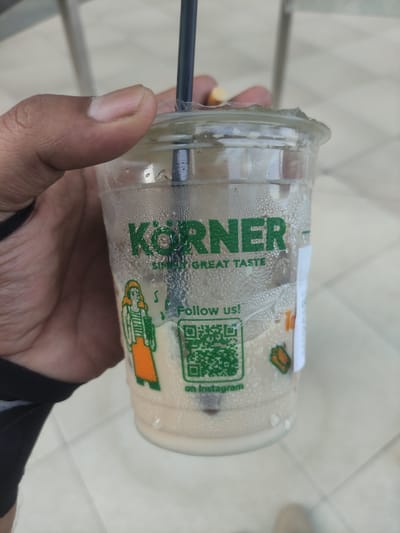 A close-up, eye-level shot captures a right hand holding a transparent plastic cup containing a cold beverage, positioned in Rawa Burung, Indonesia. The cup exhibits significant condensation droplets on its exterior, indicating the presence of a chilled liquid. A black plastic straw is inserted through a circular opening in the clear plastic lid. The beverage inside is a light beige or cream-colored liquid, with several dark brown, granular particles settled at the bottom of the cup.

The cup features green branding that prominently displays "KÖRNER" in large capital letters, followed by "SIMPLY GREAT TASTE" in a smaller font. To the left of the text, a cartoon illustration of a figure, appearing to be a woman with an orange skirt and white top, is printed on the cup. Below the "KÖRNER" text and to the right of the cartoon, the phrase "Follow us!" is printed above a square green QR code, which has "on Instagram" printed beneath it. A small, light brown, oval-shaped object is visible near the top of the straw, held or touched by a finger of the holding hand. The hand itself has a medium-dark skin tone, with the thumb and part of the index finger visible gripping the cup. A black sleeve or wristband is partially visible on the person's left wrist. The background is a soft, light-toned blur, revealing what appears to be a tiled floor pattern.