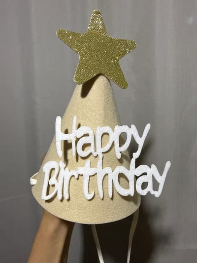 A person is holding a cream-colored birthday cone hat. The hat is decorated with a gold glitter star at the top and white felt lettering that spells "Happy Birthday." The hat has white elastic straps attached to it. The background is a plain gray fabric. The image appears to be taken indoors, with a single light source casting a shadow on the right side of the hat. There are no indications of the city of Nerudstal, Ukraine.