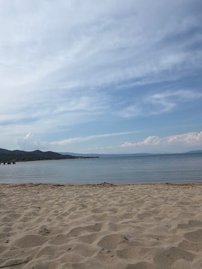 The image depicts a serene beach scene at Olympiada, Greece. The foreground is dominated by textured sand dunes, showing gentle ripples and scattered small twigs. The midground features the calm, light blue sea, with subtle horizontal waves reflecting the sky. In the distance, a chain of low, tree-covered hills forms the coastline, with a small pier or jetty extending into the water on the left side. The sky is a bright blue, partially covered by wispy white clouds that stretch across the horizon. The overall impression is one of tranquility and natural beauty.