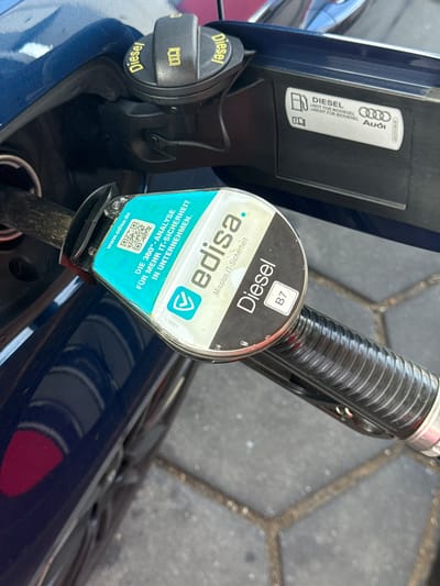 This is a close-up, eye-level shot of a diesel fuel nozzle inserted into the fuel filler of a dark blue car. The setting appears to be a gas station, suggested by the textured ground and the presence of the fuel pump.

The central focus is the fuel nozzle, with a prominent turquoise and white label featuring the "edisa." logo, the words "Mission IT-Sicherheit," and "DIE 360°-ANALYSE FÜR MEHR IT-SICHERHEIT IN UNTERNEHMEN." A QR code and the website "www.edisa.de" are also visible on the label. Above this, a small black label indicates "Diesel" and "B7".

To the left of the nozzle, the car's fuel filler cap is open and features a black cap with yellow text that also reads "Diesel" and a symbol of a fuel pump. A sticker on the car's bodywork, next to the filler neck, displays the Audi logo and the text "DIESEL >NOT FOR BIODIESEL >NICHT FÜR BIODIESEL".

The lighting suggests it is daytime, likely sunny given the sharpness of the details. No people are visible in the frame. The scene is static, with the fuel nozzle poised for use, implying a refueling operation is either about to begin or has just concluded. The overall impression is one of utility and a focus on the specific type of fuel being used.