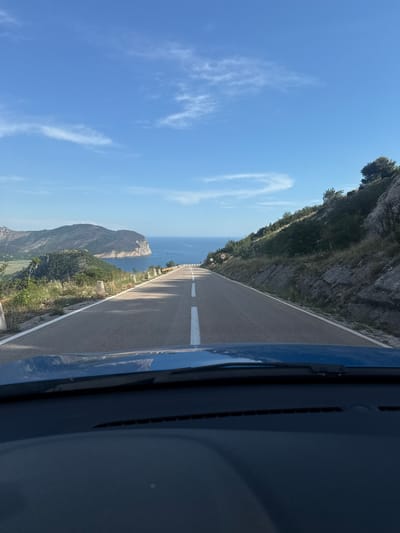 The image shows a view from a vehicle driving on a road. The road curves to the left and ascends a hill. On the right side of the road, there is a rocky, steep incline with sparse vegetation. On the left side of the road, the landscape drops away to reveal a bay with calm blue water. Mountains and hills are visible on the opposite side of the bay, with a prominent, rounded peninsula jutting out into the sea. The sky is clear blue with scattered white clouds. There are a few speed limit signs visible along the roadside. The foreground shows the dashboard and windshield of the vehicle. The scene appears to be a scenic coastal road, likely in a mountainous region.