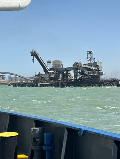 The image captures an industrial port or terminal scene under a clear, bright blue sky. In the foreground, the dark-colored hull and blue railing of a ship or large vessel are visible on the right side, extending into the bottom right corner. Some blue and yellow fittings are also seen on the vessel's exterior.
To the left of the ship, a large body of choppy, greenish-blue water extends across the middle ground. Beyond the water, an extensive industrial complex lines the shore or pier. This complex features multiple large cranes and conveyor belt systems, characteristic of bulk cargo handling operations. These towering structures, made of dark metal, are equipped with various platforms and mechanisms for moving materials. Piles of what appear to be dark bulk cargo, possibly coal or mineral ore, are visible on the dock behind these structures, suggesting active storage or processing. A faint haze or dust cloud is present above parts of the complex, hinting at ongoing operations.
The activity depicted is likely the loading or unloading of bulk materials by the industrial machinery, with the ship either docked for cargo transfer or passing by the terminal. The scene is illuminated by bright daylight, indicating clear weather conditions. No people are discernible, and there is no visible text on any of the subjects.