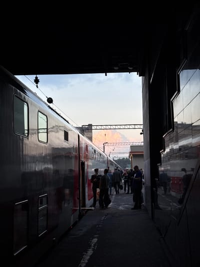 The image shows the side of a train at a station. People are standing alongside the train. The photo is taken from a dark, possibly enclosed, location, looking out towards the train and platform. The sky is visible in the distance. There is no definitive evidence to confirm the location as Ukromne, Ukraine.