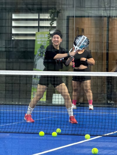 Two women are playing padel on an indoor blue court in Ban Buak Khrok North, Thailand. The woman in the foreground, on the left, is wearing a black headband, black t-shirt, and black shorts. She has on white socks and red sneakers. She is smiling and holding a padel racket in her right hand, extended towards the other player. The woman in the background, on the right, is wearing a black t-shirt and black shorts. She has on pink socks and white sneakers. She is holding a padel racket over her head. The court has a white net across the middle. Several yellow padel balls are visible on the court. In the background, there is a green banner with text that says "LEVEL UP YOUR SKILLS WITH PADEL". There is also a window with horizontal blinds, and plants hanging from the ceiling.