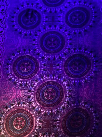 The image shows a section of a ceiling in the Ban Mae Sai Handicraft Center, located in Moo 7, Thailand. The ceiling is decorated with a pattern of circular mandalas, each containing concentric rings of geometric designs and stylized floral motifs. The color scheme is predominantly purple, with some reds and blues in the right circles. The illumination appears to be provided by purple lighting, highlighting the patterns and textures of the ceiling.