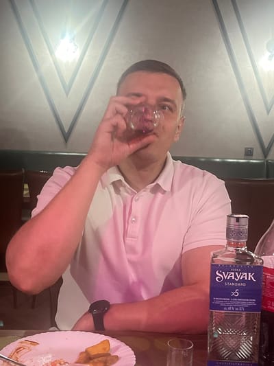 A man is holding a shot glass to his lips, about to drink. He is wearing a pink polo shirt and a black watch on his left wrist. To his right, a bottle of Svayak Standard vodka stands on a table. The bottle's label indicates it is 40% alcohol by volume. In the foreground to the right, a plate with food remnants is visible. The background shows a wall with a patterned texture and decorative lighting, suggesting an indoor setting, possibly a restaurant or bar. The image is shot from a low angle, with the man's face partially obscured by his hand and the glass. The lighting appears to be artificial, indicating it is likely evening or night. The overall mood seems casual and social. Visible text includes "VODKA SVAYAK STANDARD x5" and "alc. 40% vol. 071".